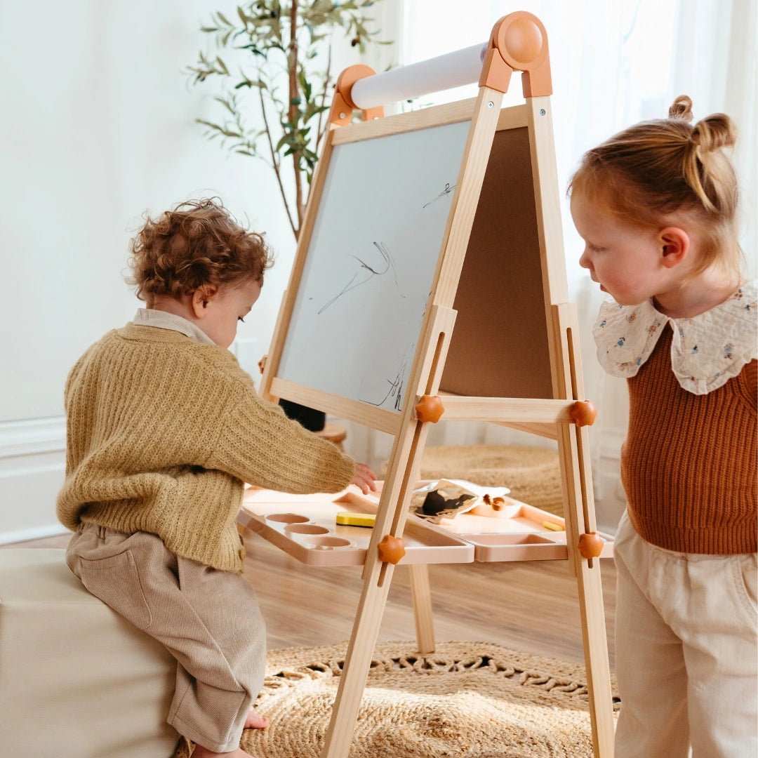 Two young children use the Tiny Land® Caramel Magnitales Easel for Kids in a cozy, sunlit room. One draws while the other watches, surrounded by light decor and a plant in the background.