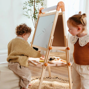 Two young children use the Tiny Land® Caramel Magnitales Easel for Kids in a cozy, sunlit room. One draws while the other watches, surrounded by light decor and a plant in the background.