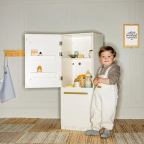 A toddler in a hoodie and white overalls stands by the Tiny Land® Play Kitchen Accessories – Fridge, a wooden cabinet refrigerator for kids. The room features neutral walls, a framed quote, and a hanging apron. Toy food and dishes are also visible.
