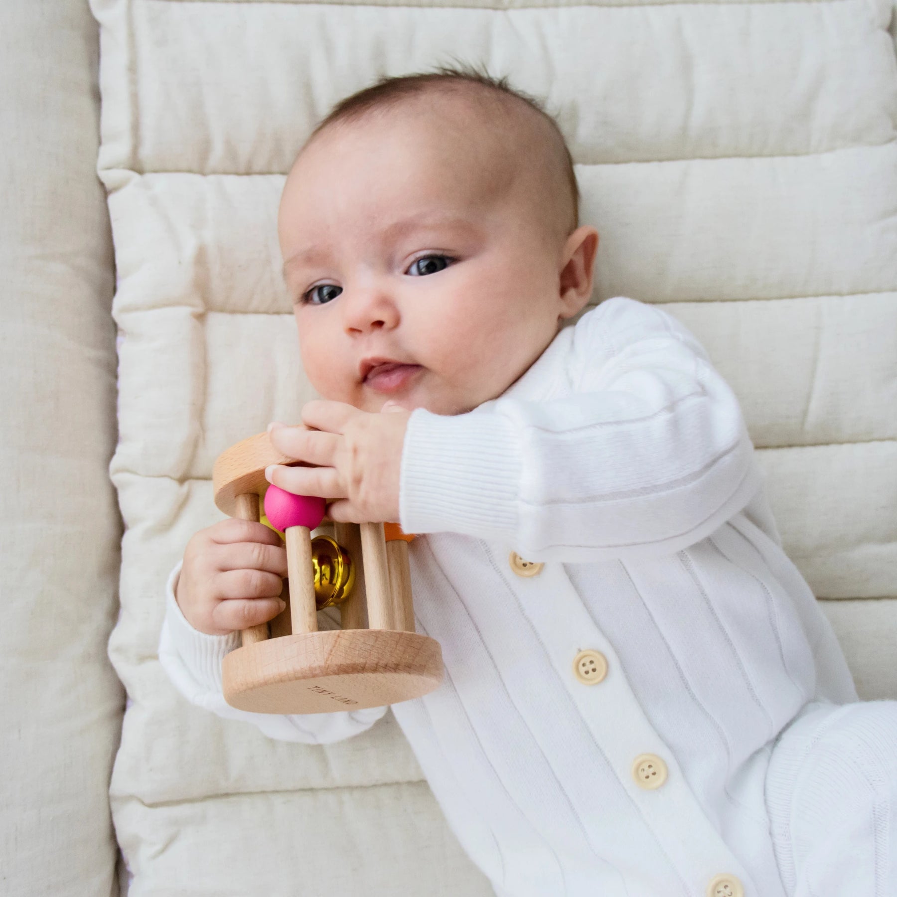 A newborn enjoys tummy time on a cream quilt, holding a Tiny Land® Montessori Toy—a wooden rattle with gold and pink details—by Tiny Land, promoting gentle sensory play for babies 0–3 months.