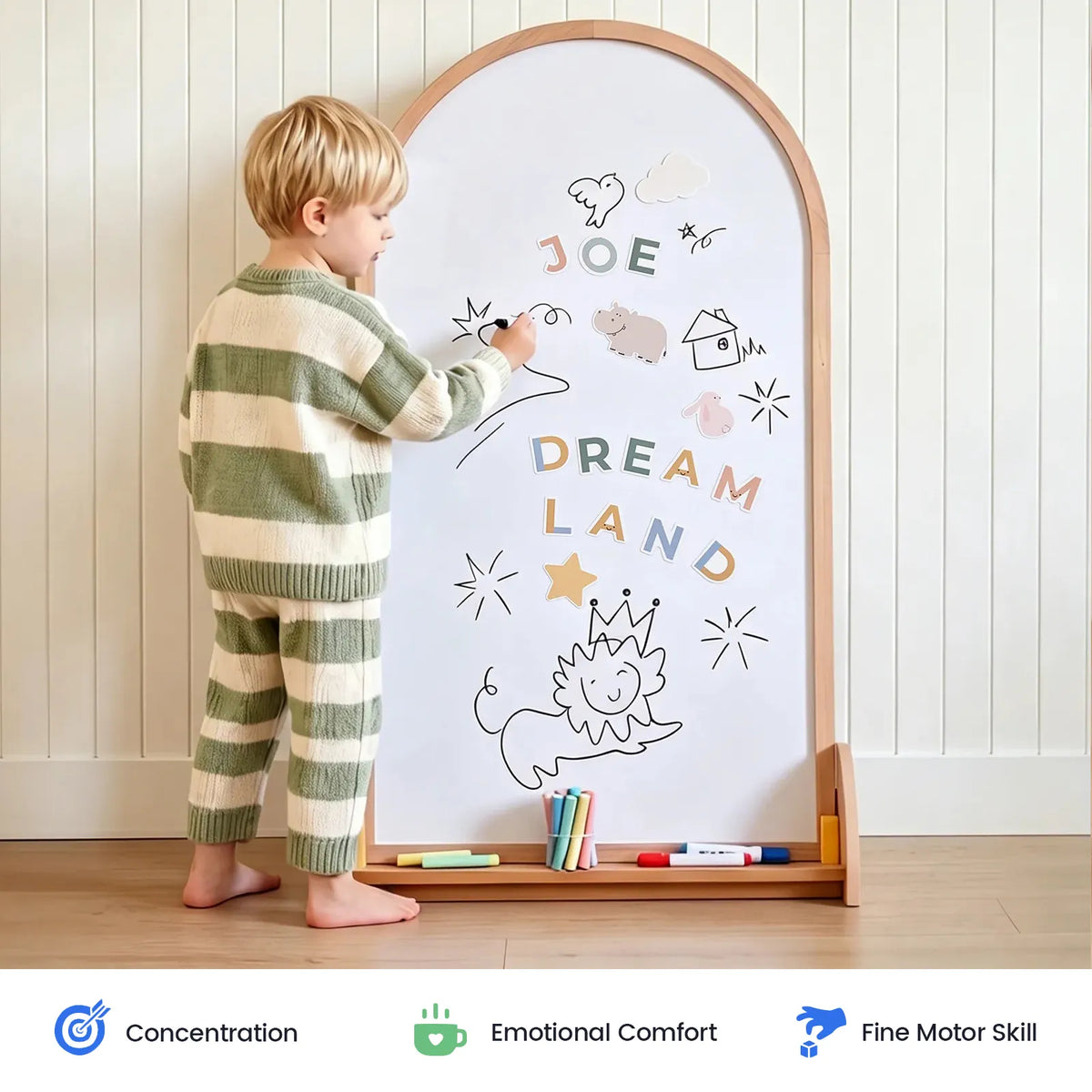 A young child in striped pajamas creates colorful doodles and writes “JOE DREAM LAND” on the Tiny Land® Double-Sided Arched Magnet Chalkboard, using vibrant markers. The playful scene showcases their concentration, emotional comfort, and fine motor skills, highlighted by icons below.