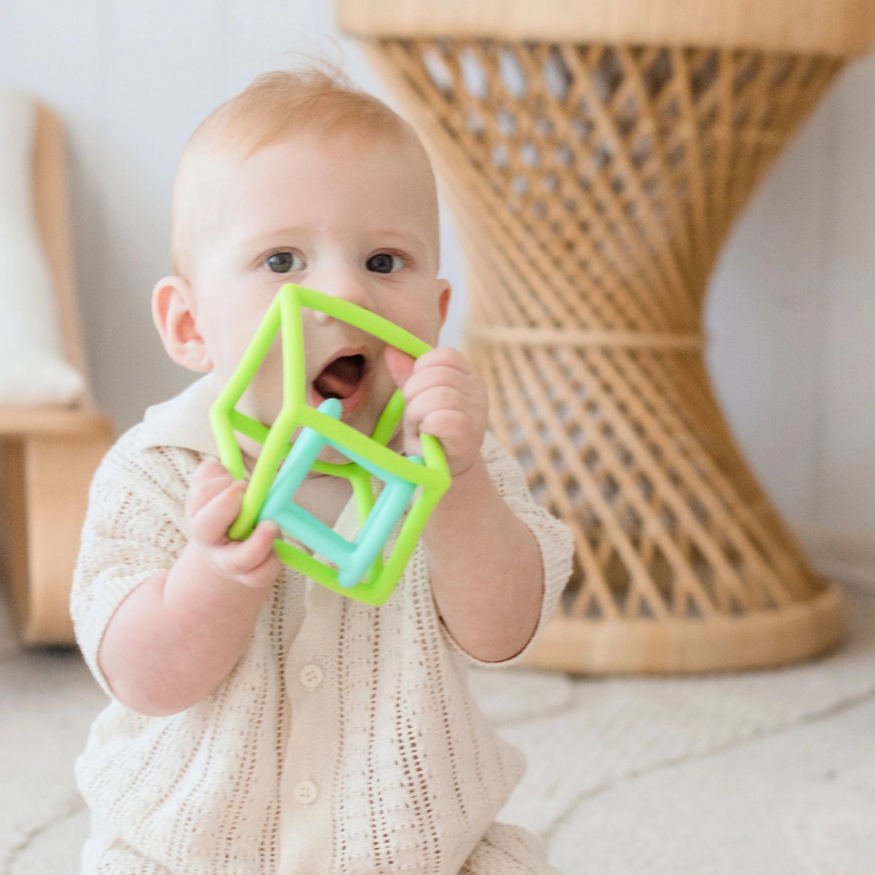 A baby in a cream outfit sits on a light rug, chewing on Tiny Land® Montessori Toys for Newborns (4–6 Months), a green and blue sensory toy that supports fine motor skill development. A wicker table is seen in the background.