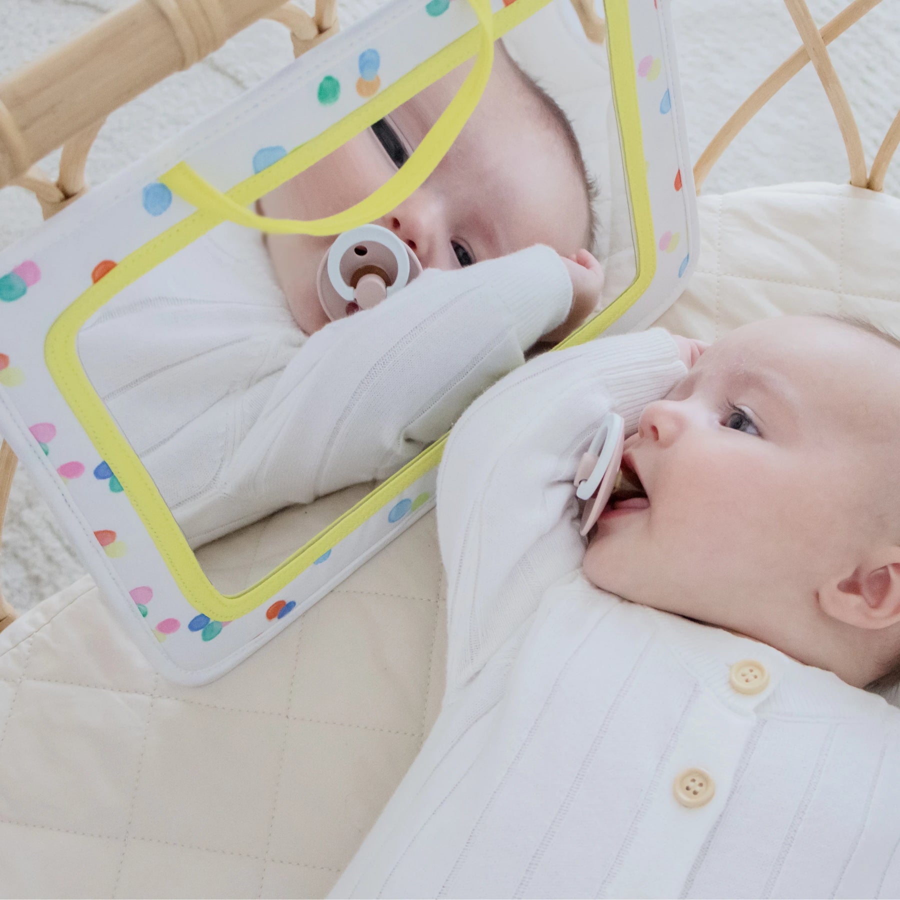 A baby in a white onesie enjoys tummy time on a quilted mat, holding a pacifier and gazing at their reflection in the colorful, rectangular Tiny Land® Montessori Toys for Newborns (0–3 Months) baby-safe sensory mirror.