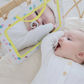 A baby in a white onesie enjoys tummy time on a quilted mat, holding a pacifier and gazing at their reflection in the colorful, rectangular Tiny Land® Montessori Toys for Newborns (0–3 Months) baby-safe sensory mirror.