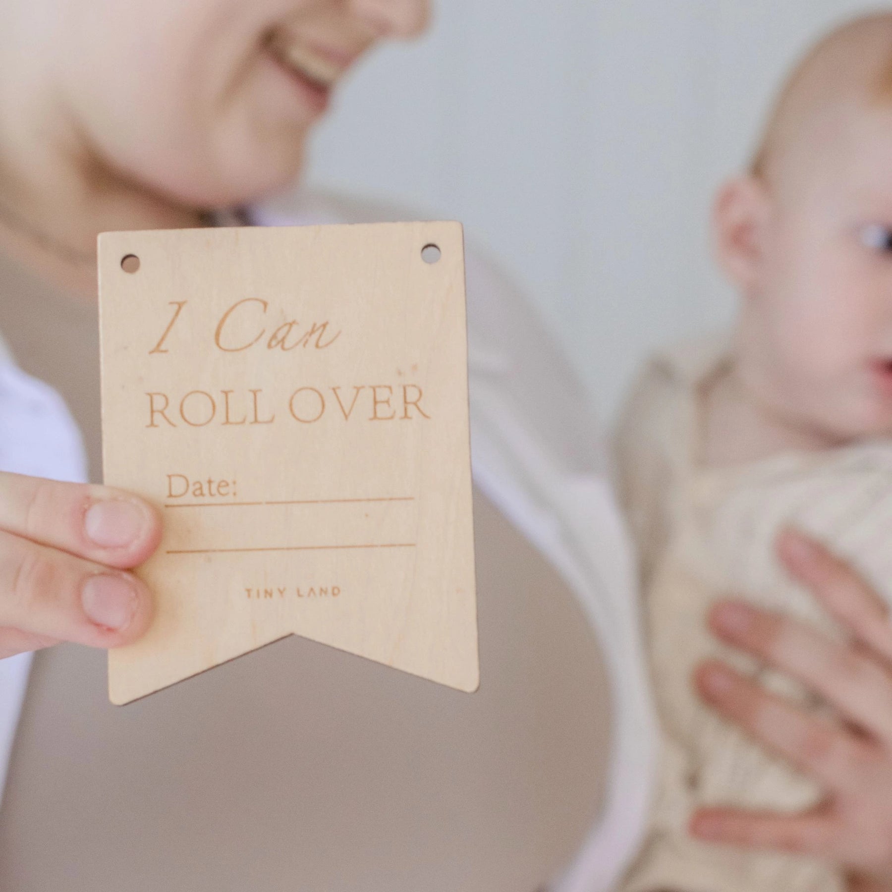 A smiling woman holds a baby and the Tiny Land® Montessori Toys for Newborns (4–6 Months) with a wooden "I Can Roll Over" plaque, perfect for milestone celebrations and nurturing fine motor skills by Tiny Land.