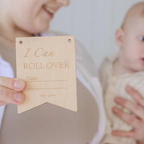 A smiling woman holds a baby and the Tiny Land® Montessori Toys for Newborns (4–6 Months) with a wooden "I Can Roll Over" plaque, perfect for milestone celebrations and nurturing fine motor skills by Tiny Land.