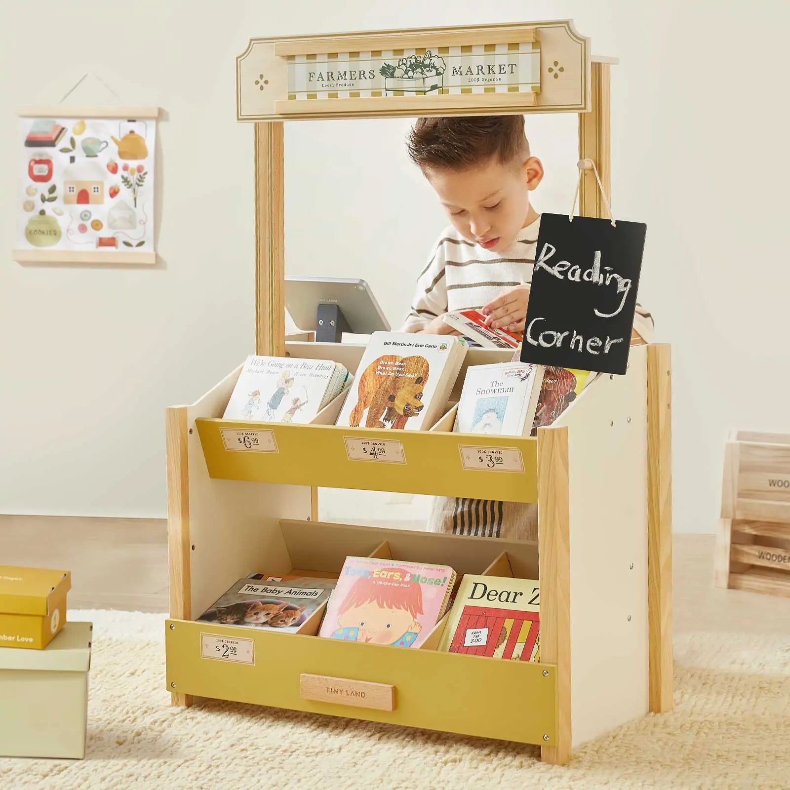 A young boy stands behind the Tiny Land® Kid's Grocery Store Playset, arranging children's books. This Tiny Land toy encourages pretend play with a "Reading Corner" chalkboard sign and more books on the lower shelves.