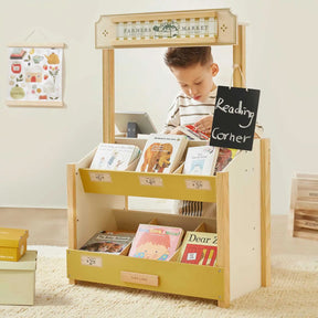 A young boy stands behind the Tiny Land® Kid's Grocery Store Playset, arranging children's books. This Tiny Land toy encourages pretend play with a "Reading Corner" chalkboard sign and more books on the lower shelves.
