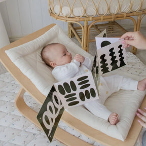 A baby in a white onesie lies in a cushioned wooden rocker, enjoying tummy time while gazing at high-contrast cards from the Tiny Land® Montessori Toys for Newborns (0–3 Months) sensory set, in a cozy, sunlit room.