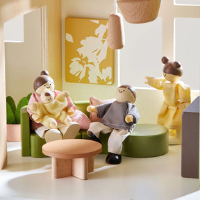Three wooden dolls relax in the Tiny Land® Sweetwood Cottage Dollhouse’s modern living room, featuring green and beige furniture, round tables, potted plants, abstract art, and bright windows for a welcoming feel.|Green