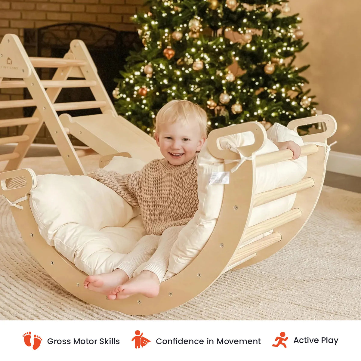 A smiling toddler sits in a cushioned wooden rocker near a decorated Christmas tree, with the Tiny Land® Thick Padded Play Cushion (arch not included) and an eco-friendly baby playmat visible beside a wooden climbing frame in the background. Icons below showcase benefits such as gross motor skill development, confidence in movement, and active play—brought to you by Tiny Land.