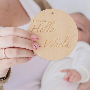 A person holds a round wooden sign reading "Tiny Land Hello World" near a newborn in white, featuring Tiny Land® Montessori Toys for Newborns (0–3 Months). The individual's nails have a pink and white wavy design.