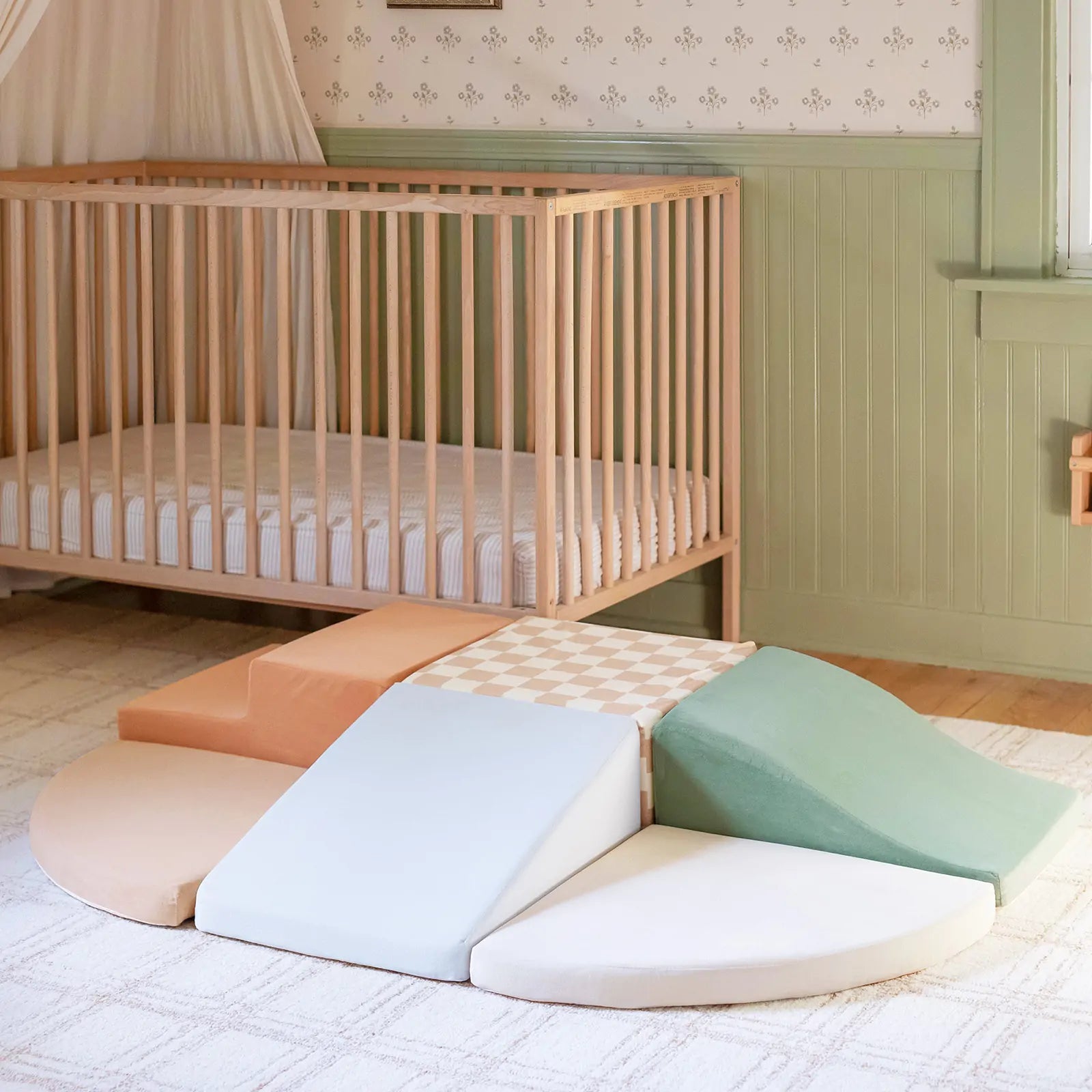 A wooden crib stands in a softly lit nursery with pale green wainscoting. On the floor, Tiny Land® PlayNest Climbing Blocks by Tiny Land, in pastel peach, green, cream, and checkered beige, encourage gross motor development.