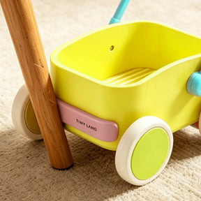 A close-up of the Tiny Land® 3-in-1 Wooden Baby Walker Wagon - Spring Color, featuring a yellow body, green wheels, and a blue handle, sits on a beige carpet beside a wooden storage box and furniture leg.