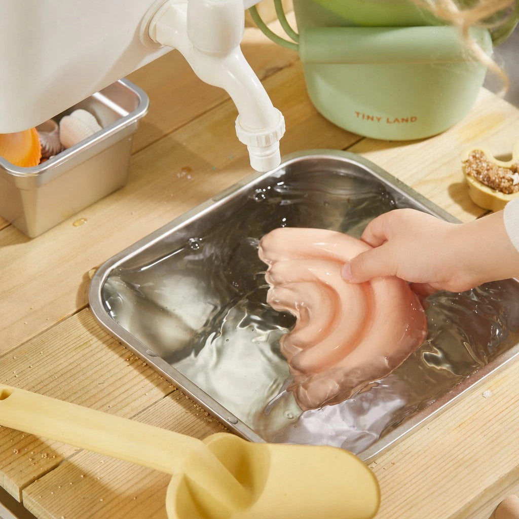 A child washes the soft, pink, rainbow-shaped sand toys from the Tiny Land® Outdoor Mud Kitchen & Sand Play Set in a metal tray of water. Nearby are a plastic scoop, kids’ kitchen utensils, and a green cup—ideal for sensory outdoor fun.