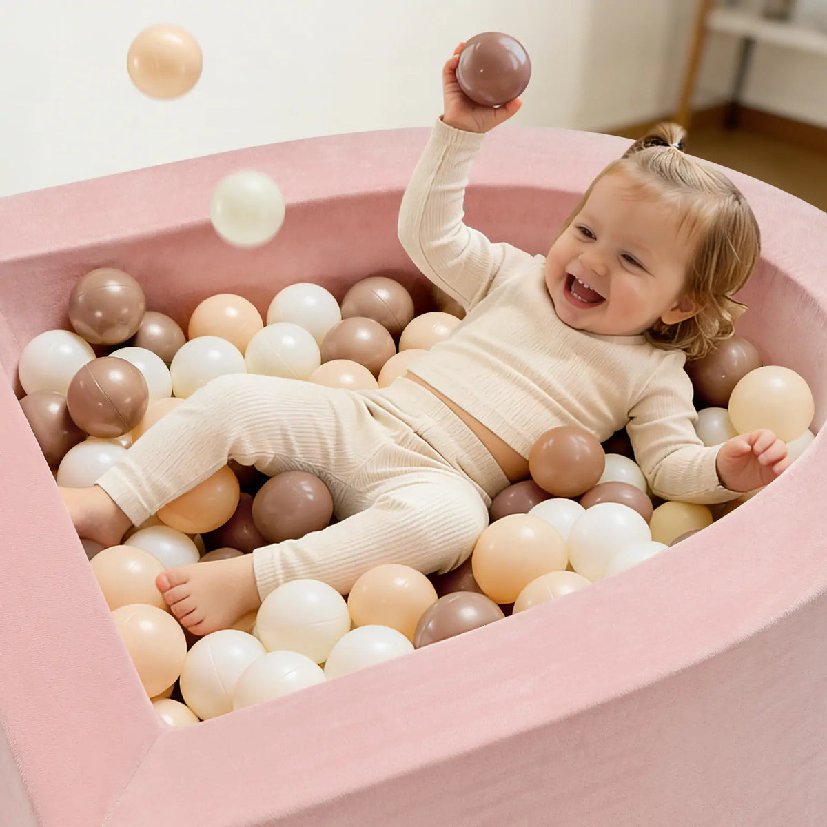 A smiling toddler in cream clothes plays in the Tiny Land® PlayNest Climbing & Ball Pit Set by Tiny Land, tossing a ball while enjoying sensory fun amidst beige, white, and brown balls in the pink, heart-shaped foam pit.