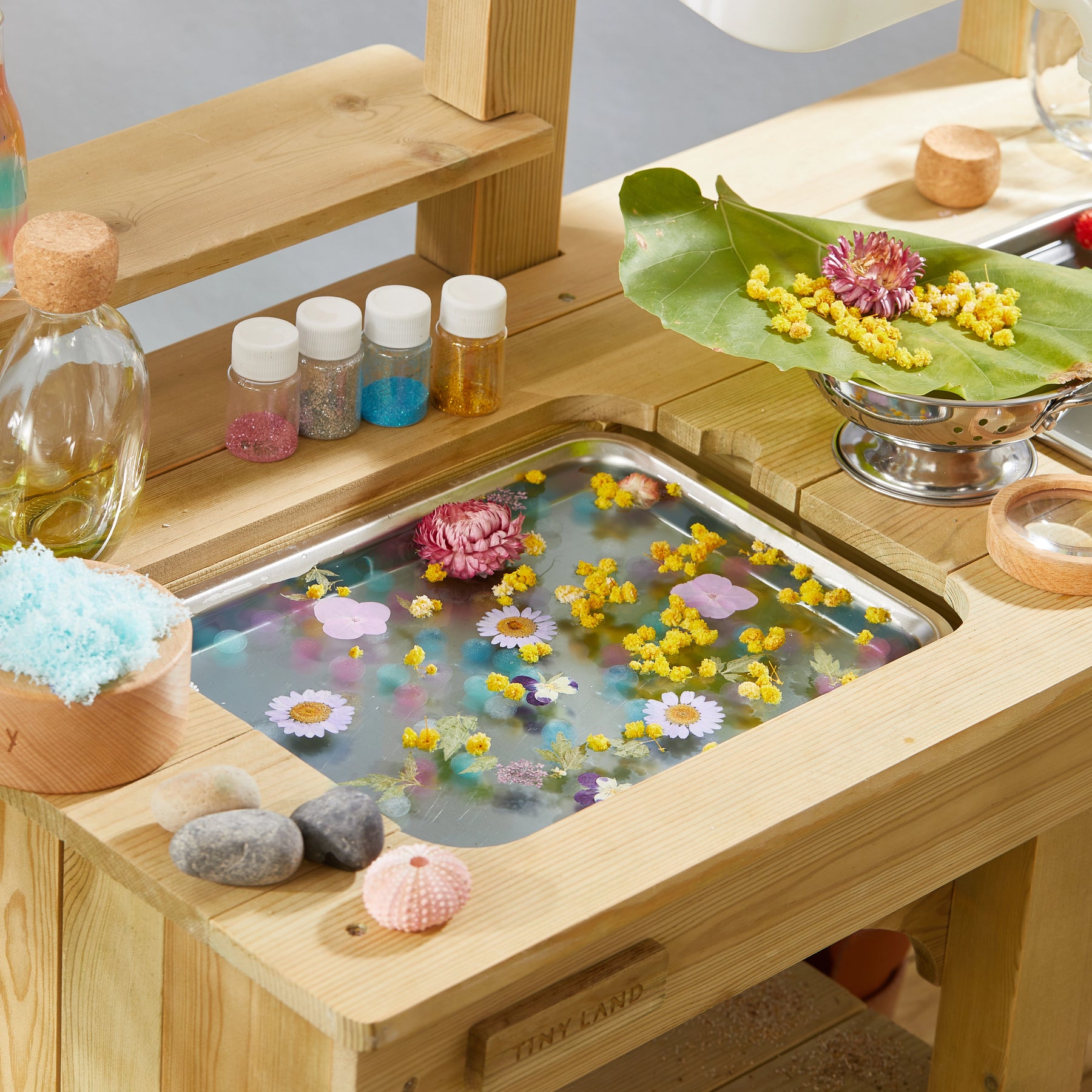 The Tiny Land® Outdoor Mud Kitchen & Sand Play Set by Tiny Land features a wooden sink with water, floating flowers, petals, glitter bottles, a green leaf with flowers, pink seashell, stones, blue bath salt, and a silver strainer for outdoor play.
