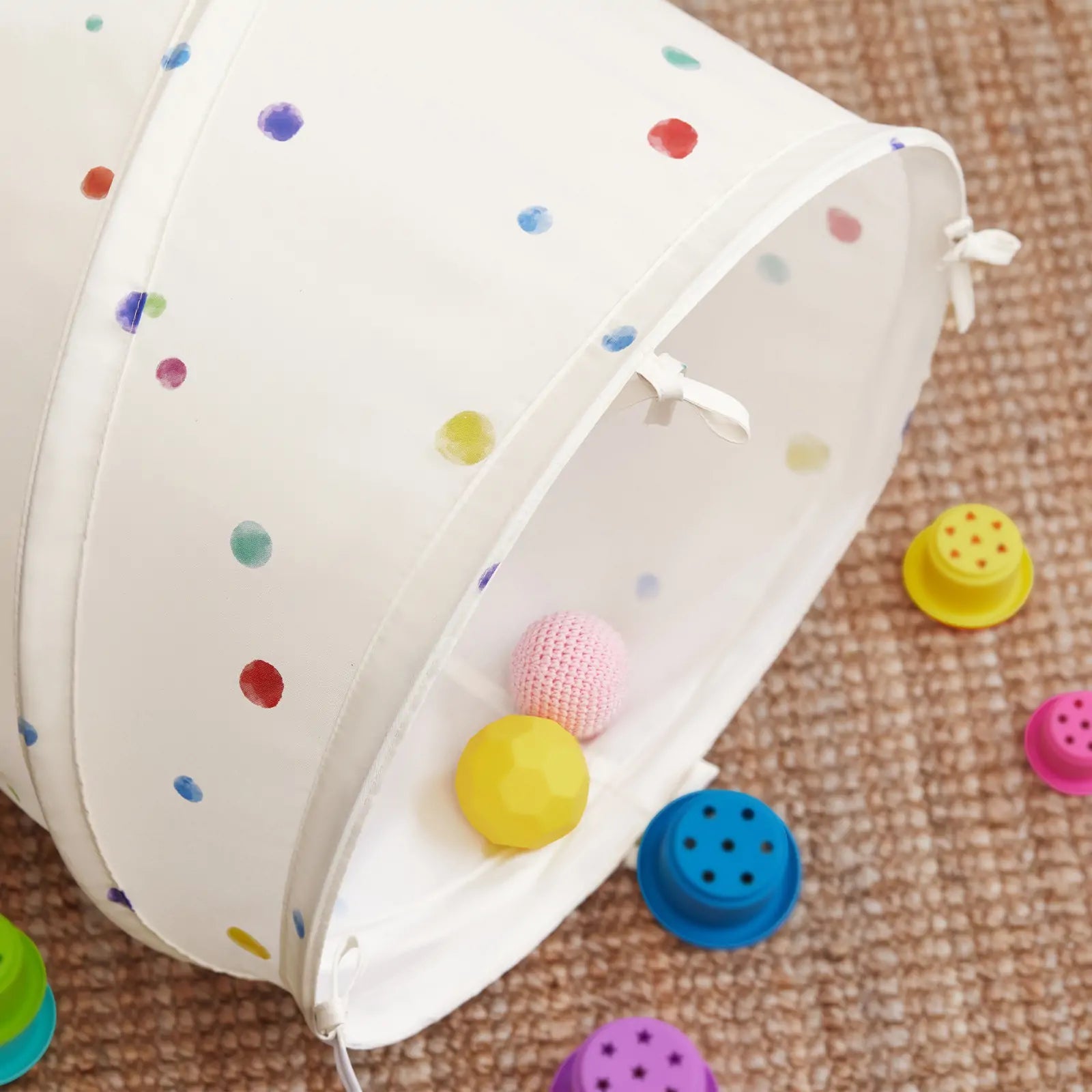 The Tiny Land® Adventure Play Tunnel - Colorful Polka Dots by Tiny Land rests on a woven carpet, with textured balls and stacking cups scattered around its opening, creating an inviting space for kids' active play.