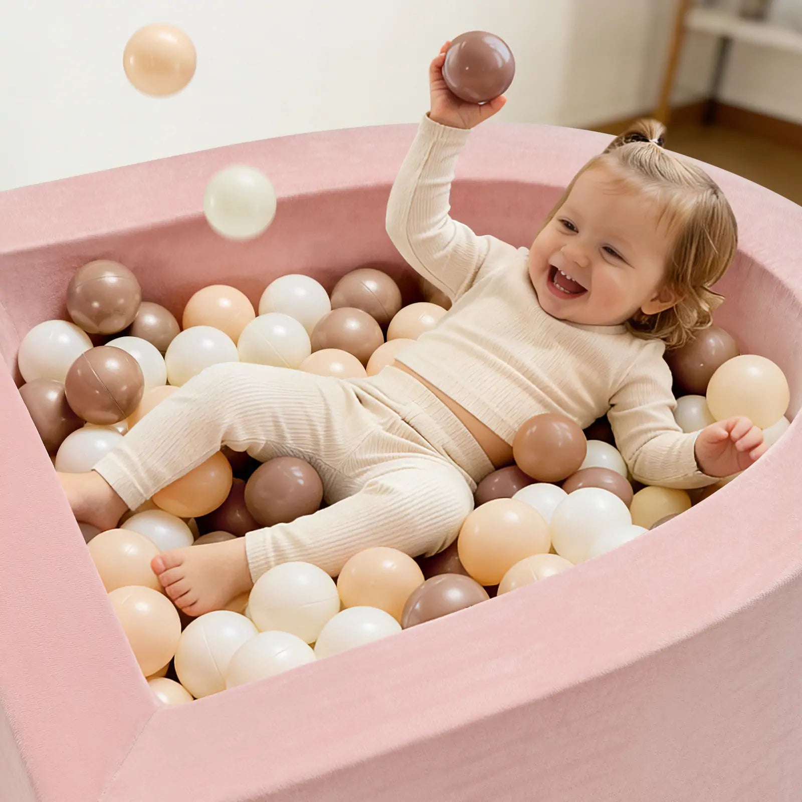 A joyful toddler dressed in beige enjoys playtime in the Tiny Land® PlayNest Climbing & Ball Pit Set—a soft, pink, heart-shaped foam ball pit by Tiny Land—filled with white, beige, and brown balls and holding one up happily in this cozy indoor playground.|Pink