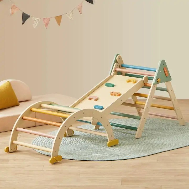 A cozy room showcases the Tiny Land® 7-in-1 Morandi Pikler Triangle Set by Tiny Land, featuring a wooden arch, triangle, slide with handholds, colorful rungs—set atop a round rug with a cushion and festive wall bunting in the background.|morandi color