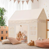 Two children enjoy the Tiny Land Bright Play & Art Set, featuring a colorful easel and a cream playhouse with lights, surrounded by pillows and toys in a bright, plant-filled room—perfect for imaginative indoor or outdoor fun|white