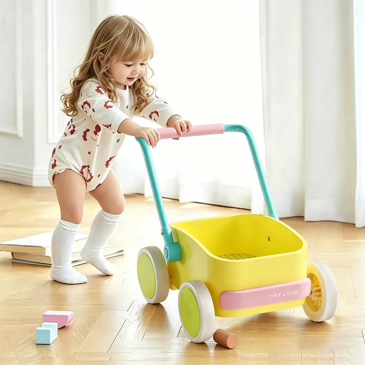 A young child in a patterned onesie and knee-high socks pushes the Tiny Land® 3-in-1 Wooden Baby Walker Wagon - Spring Color indoors on a wooden floor, with blocks nearby and adjustable speed control for safety.