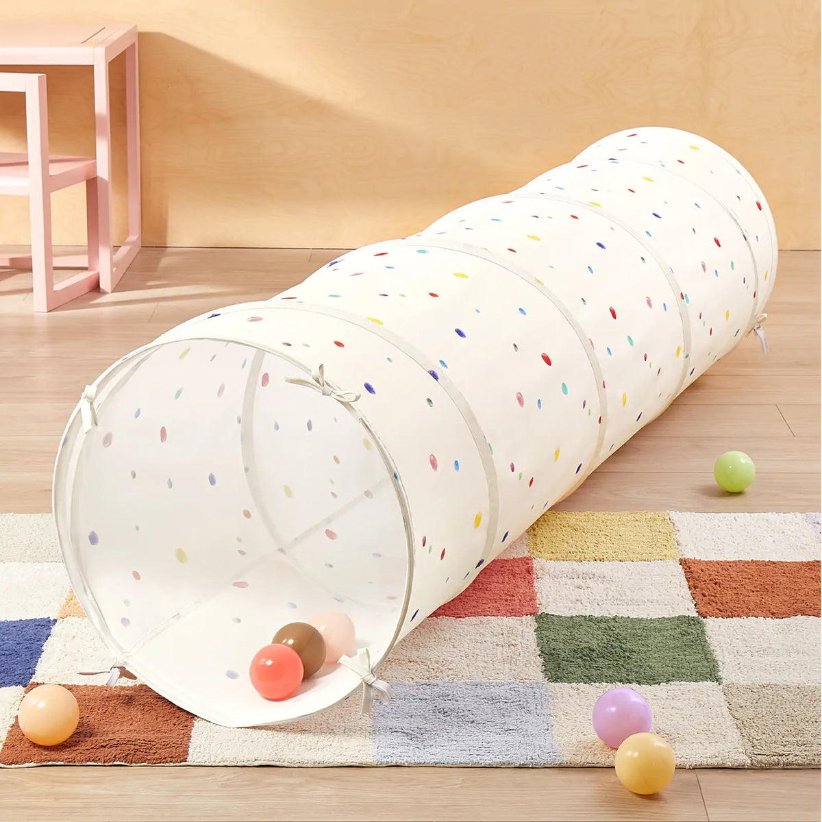 The Tiny Land® Adventure Play Tunnel by Tiny Land, featuring a white design with colorful polka dots, sits on a checkered rug and is surrounded by scattered plastic balls in a bright, child-friendly room.|Colorful Dots