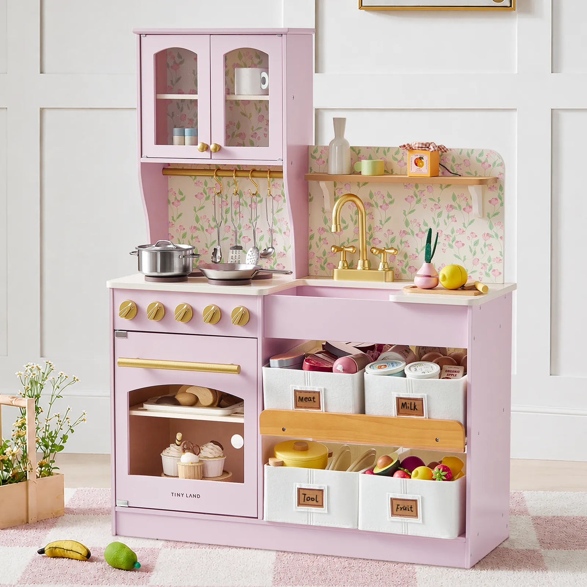 The Tiny Land® Wooden Play Kitchen with Storage for Creative Toddlers by Tiny Land comes in pink with gold accents and includes pretend cookware, toy food, pots, and labeled storage bins for meat, milk, tools, and fruit. Set against floral wallpaper for added charm, this multifunctional play kitchen features a sink, oven, stove, and thoughtful storage—while decorative flowers placed nearby complete the inviting scene.|Sakura Pink