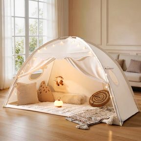 The Tiny Land® Kids Bed Tent by Tiny Land features white fabric, string lights, plush pillows, a stuffed toy, and a soft mat. It stands on a wooden floor in a bright living room with large windows letting in natural light.|Beige
