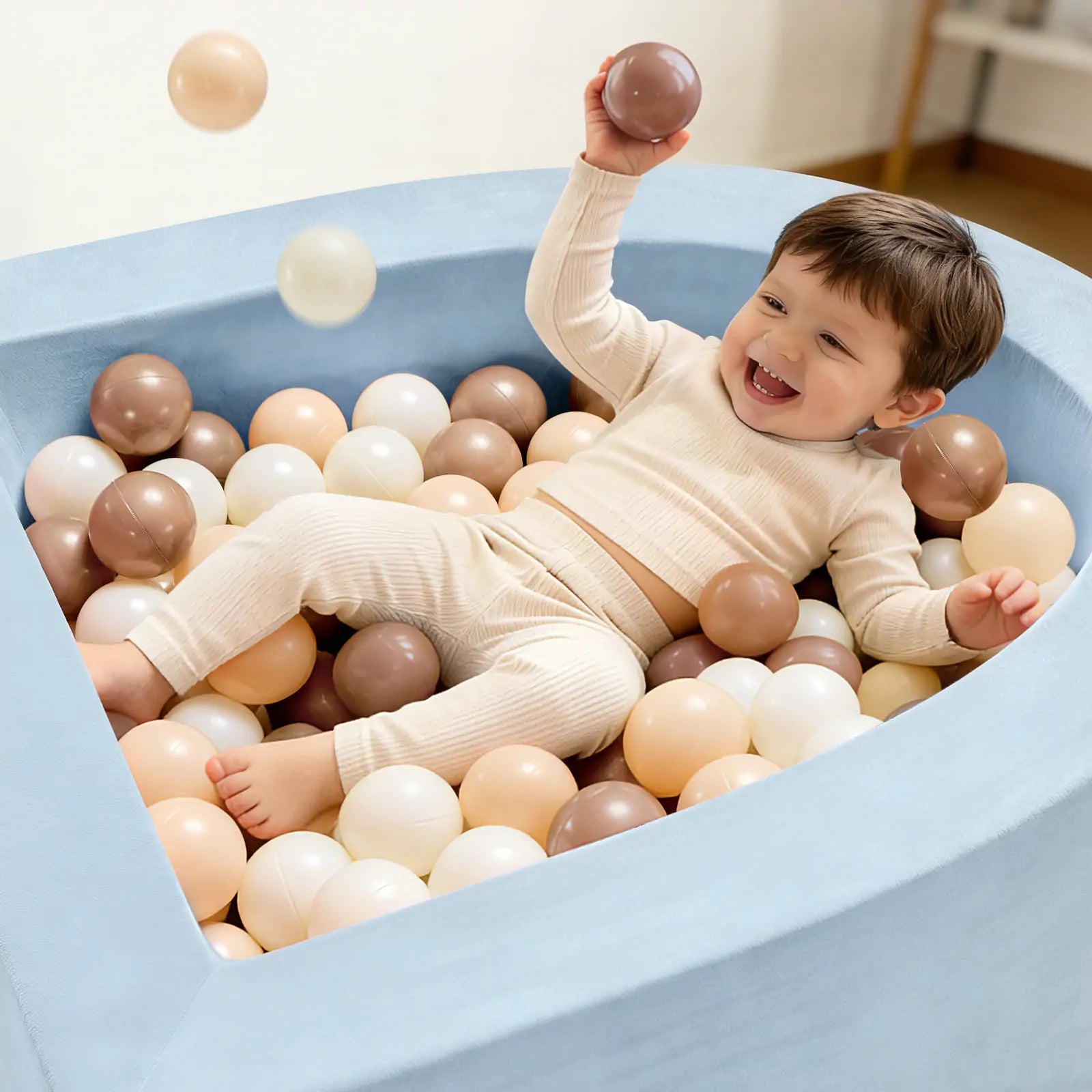 A smiling toddler in cream-colored pajamas plays in the Tiny Land® Violet PlayNest Climbing Ball Pit Set with Tunnel, filled with beige, brown, and white balls, at an indoor playground—joyfully tossing one into the air.|Blue