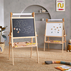 Two Tiny Land® Caramel Magnitales Easels for Kids are in a playroom—one features a chalkboard with "ABC" and "IDEAS"; the other is magnetic with colorful letters. Art supplies and toys fill the shelves and floor.