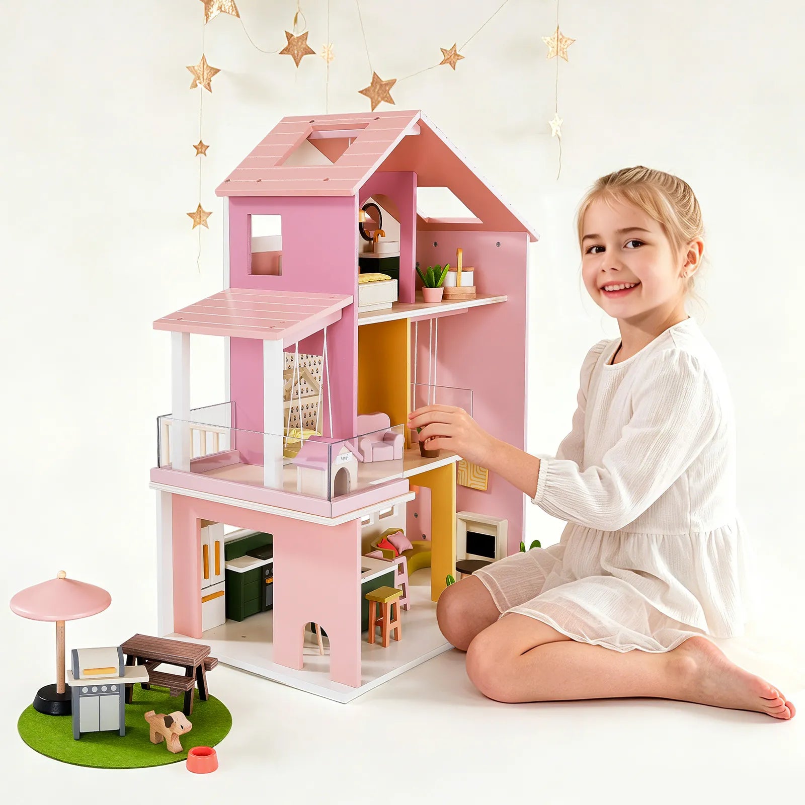 A young girl smiles as she plays with the Tiny Land® Sweetwood Cottage Dollhouse, a pink and white wooden set with multiple rooms and furniture, plus a small outdoor playset. Gold star decorations hang in the background.|Pink