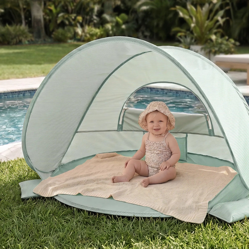 A smiling baby in a sun hat sits on a blanket inside the Tiny Land® 4-in-1 Baby Beach Tent & Sand Toy Set by Tiny Land, ready for sand and water play by the pool, surrounded by grass and greenery.