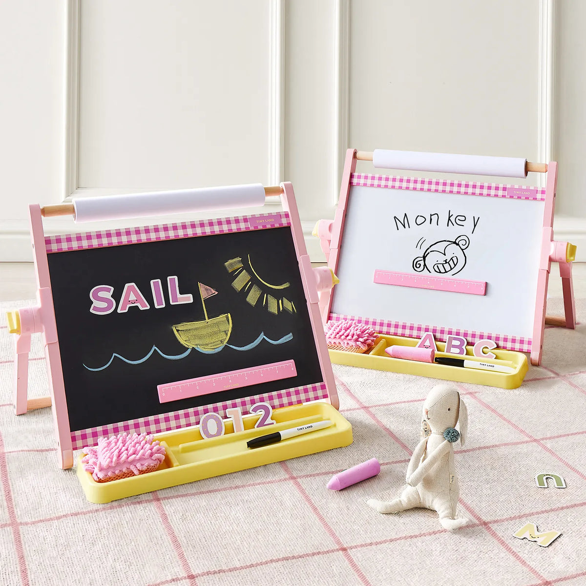 Two pink-framed Tiny Land® Double-Sided Magnetic Tabletop Easels by Tiny Land—each serving as a vibrant kids' art station—display drawings: one featuring a sun, sailboat, and the word "SAIL" on its magnetic whiteboard, the other showing a monkey face with "Monkey." A plush toy and alphabet cards rest on the carpeted floor in front.|Pink