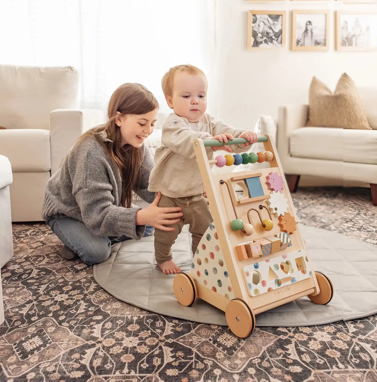 A woman smiles as she supports a toddler standing with the Tiny Land® Versatile Natural Wooden Baby Walker on a colorful patterned rug in a cozy living room.