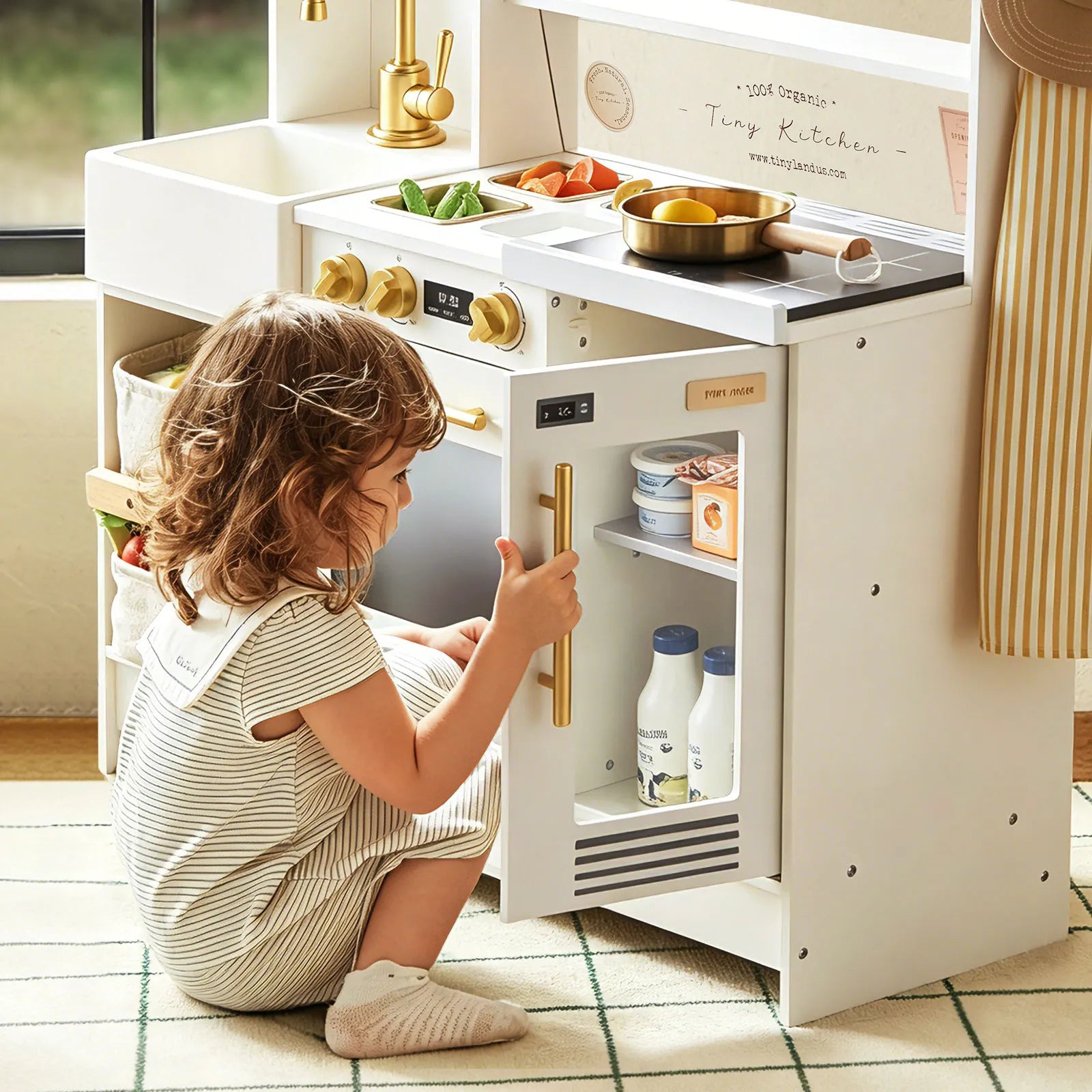 A young child in a striped dress sits on a rug, enjoying pretend play with the Tiny Land® BistroSwitch Play Kitchen, opening the toy fridge to explore play food and bottles inside.|White