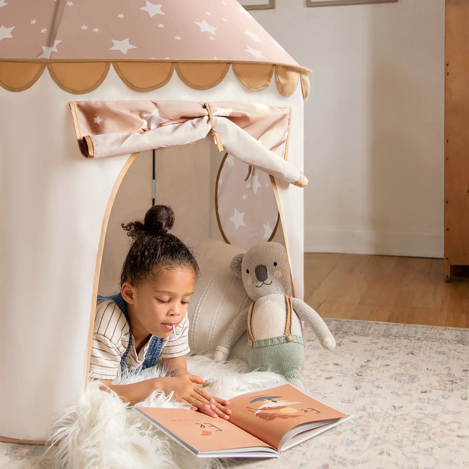A young child reads a book inside the Tiny Land® Kids Tent – Indoor Tent for Toddlers by Tiny Land, with a stuffed koala nearby on a soft rug, creating a cozy and playful atmosphere in the softly lit room.|Brown