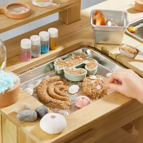 A child shapes sand in a metal tray with molds and seashells from the Tiny Land® Outdoor Mud Kitchen & Sand Play Set by Tiny Land, surrounded by jars of colored sand, stones, and creative materials on a wooden table.
