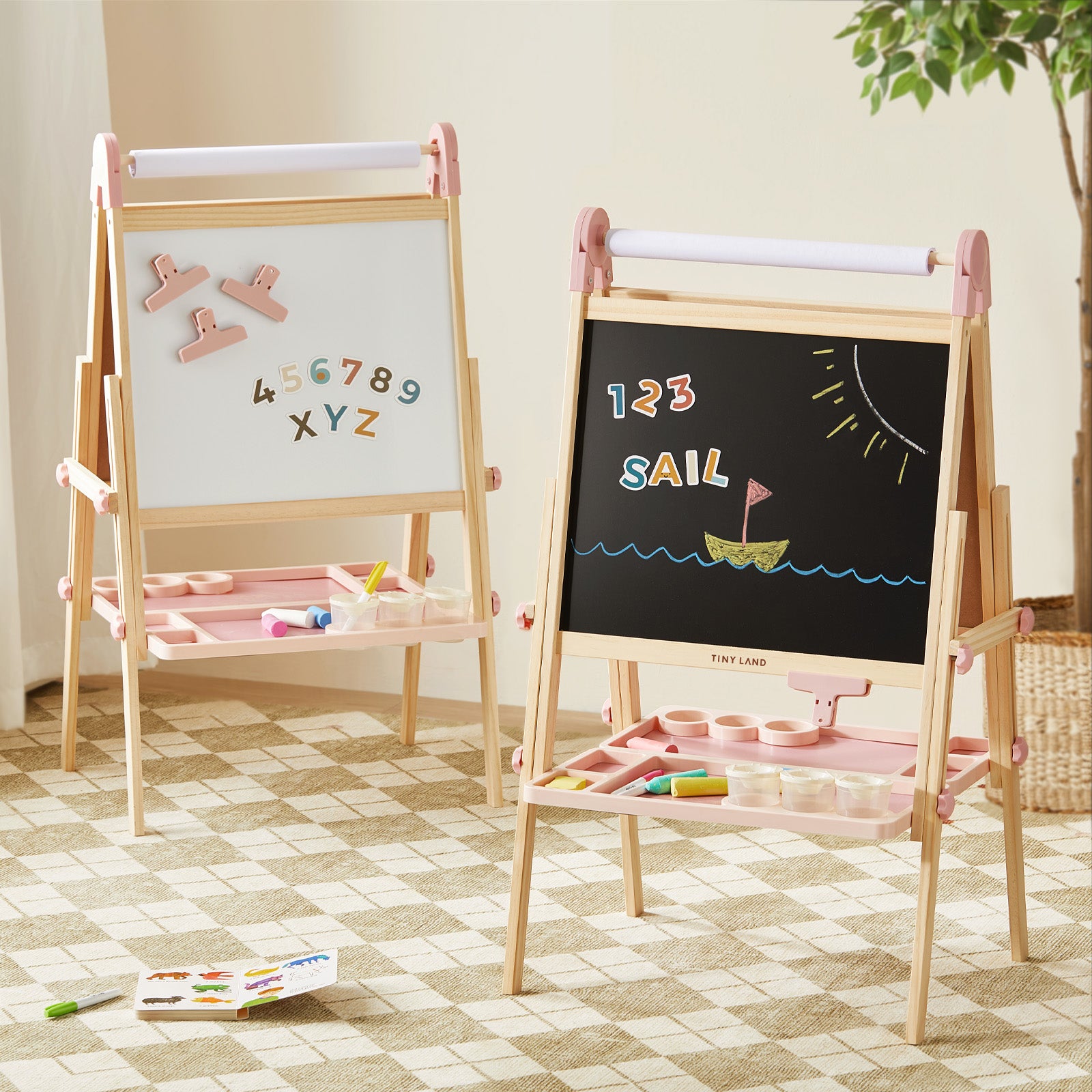 Two Tiny Land® Magnitales Easel for Kids - Pink by Tiny Land stand on a checkered rug: one displays magnetic letters and numbers, the other shows “123 SAIL” with a sailboat. Art supplies sit on trays beneath each easel.|pink