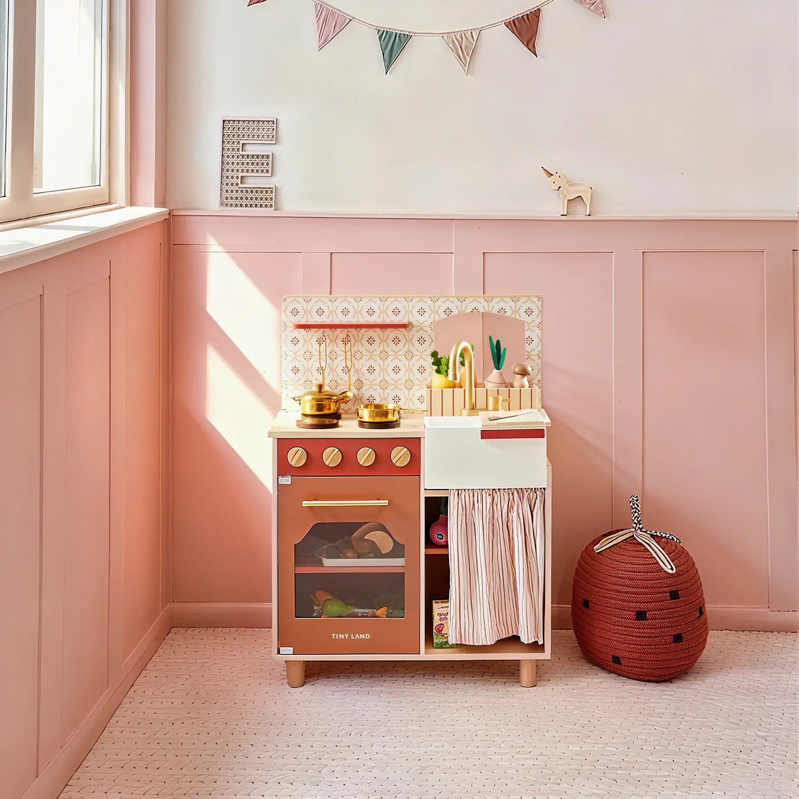 The Tiny Land® Mediterranean Play Kitchen by Tiny Land, in pink and gold, sits in a pastel pink room with a window, bunting, soft flooring, a red strawberry-shaped storage basket, and a small giraffe figurine nearby|Mediterranean