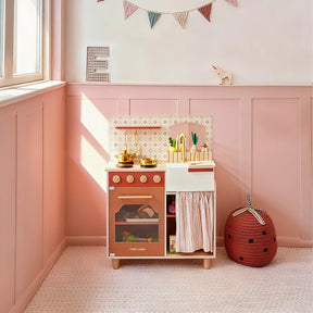 The Tiny Land® Mediterranean Play Kitchen by Tiny Land, in pink and gold, sits in a pastel pink room with a window, bunting, soft flooring, a red strawberry-shaped storage basket, and a small giraffe figurine nearby|Mediterranean