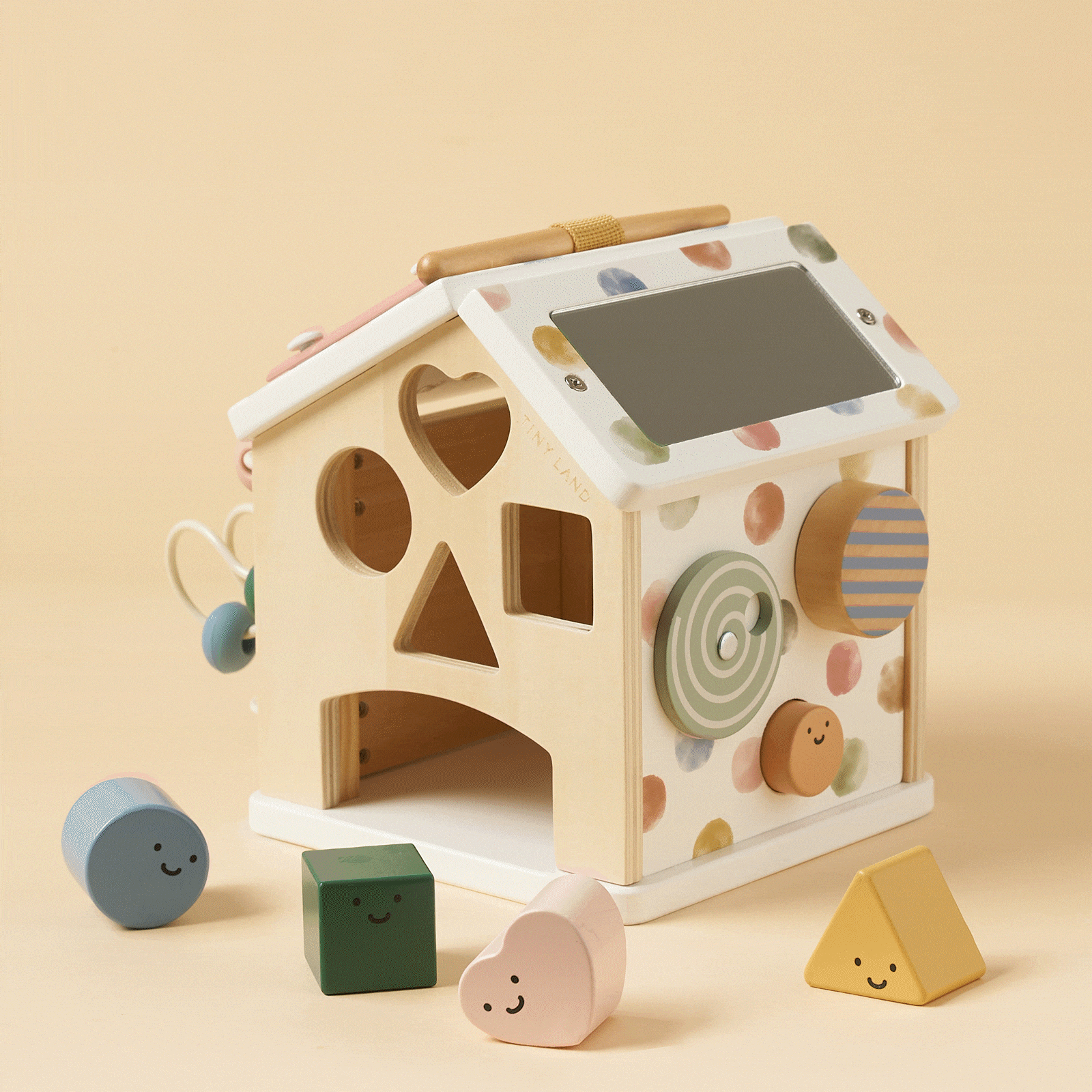 The Tiny Land Cozy Learning Playset includes a playmat and pastel activity cube with cut-out circle, triangle, square, and heart shapes. Smiling blocks are scattered around the toy on a beige background.