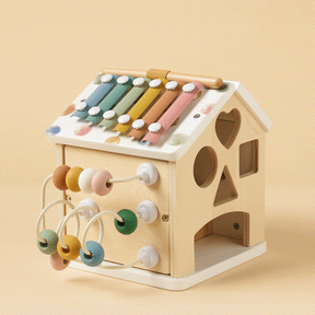 The Tiny Land Cozy Learning Playset features a wooden house-shaped activity cube with a colorful xylophone roof, mallet, pastel abacus, and shape cutouts on a soft beige playmat—perfect for engaging developmental play.
