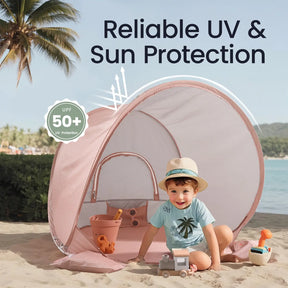 A young child in a sun hat sits inside the Tiny Land® One-Stop Beach Toy & Tent Set – Blush Pink by Tiny Land on sand, playing with a sand toy set. The UPF 50+ tent features “Reliable UV & Sun Protection.” Palm trees and water appear in the background.|Blush Pink
