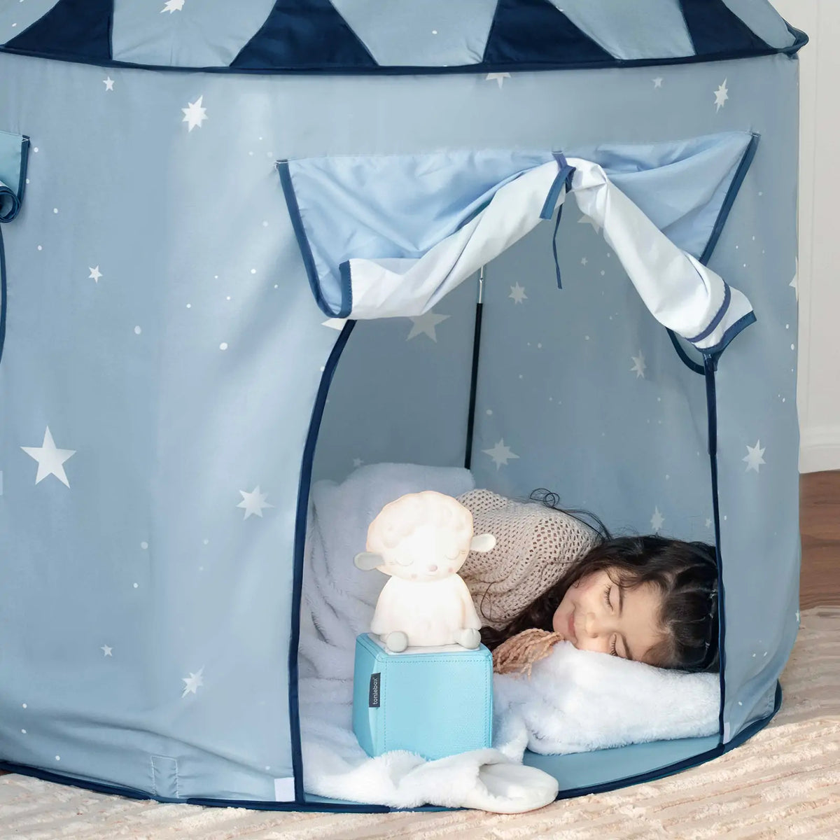 A young child sleeps in the Tiny Land® Kids Tent – Indoor Tent for Toddlers by Tiny Land, resting on a white pillow and blanket. A glowing plush toy sits near her head inside the cozy blue tent with star patterns and open curtains.|Blue