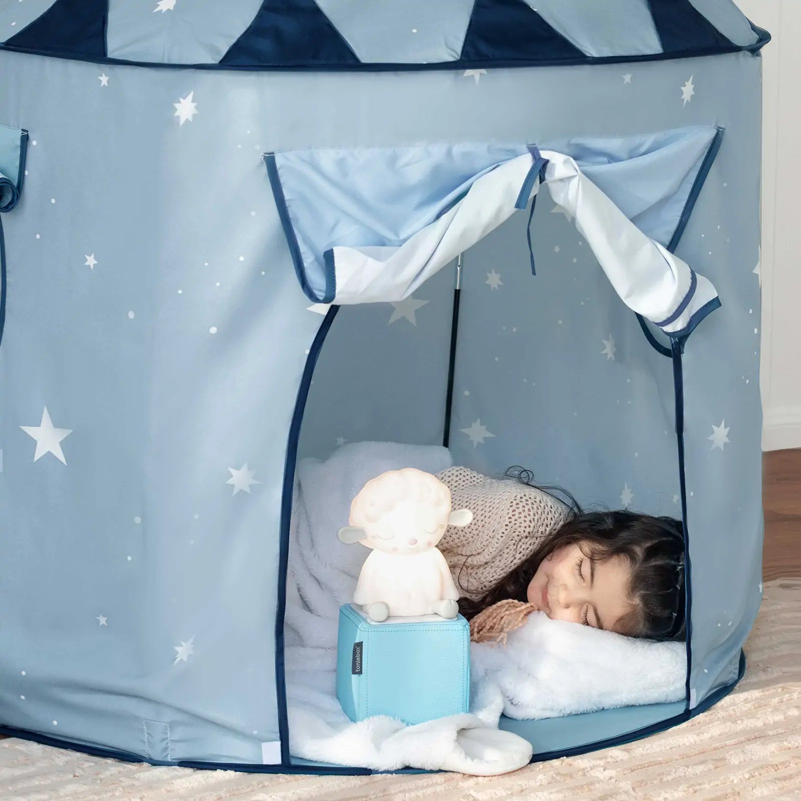 A young child sleeps in the Tiny Land® Kids Tent – Indoor Tent for Toddlers by Tiny Land, resting on a white pillow and blanket. A glowing plush toy sits near her head inside the cozy blue tent with star patterns and open curtains.|Blue