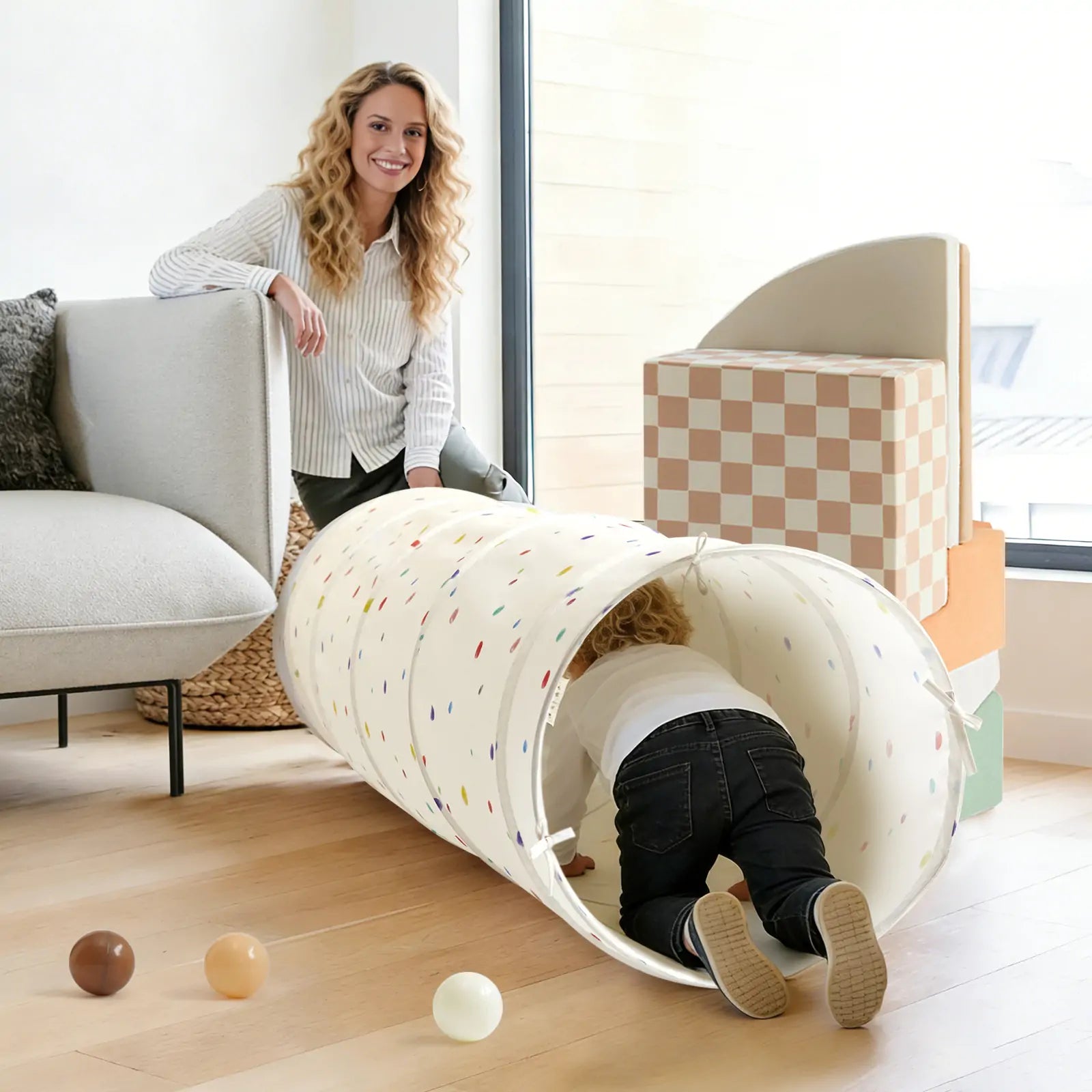 A woman smiles on a couch as a child crawls through the Tiny Land® Adventure Play Tunnel - Colorful Polka Dots. Soft play blocks and balls from Tiny Land are scattered on the wooden floor in this bright, cheerful room.