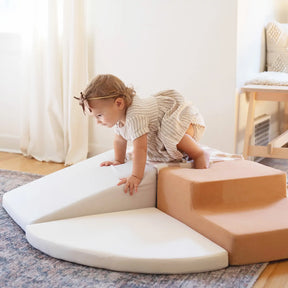 A toddler in a light dress and bow climbs on Tiny Land® PlayNest Climbing Blocks by Tiny Land in a cozy, sunlit room with a rug and wooden floor, encouraging gross motor development.