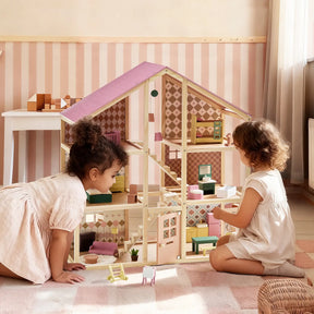 Two young children play on the floor with the Tiny Land® Sweetwood Pegasus Dollhouse & Wooden Dollhouse Dolls. The room’s pink stripes and pastel decor add a warm, playful atmosphere.