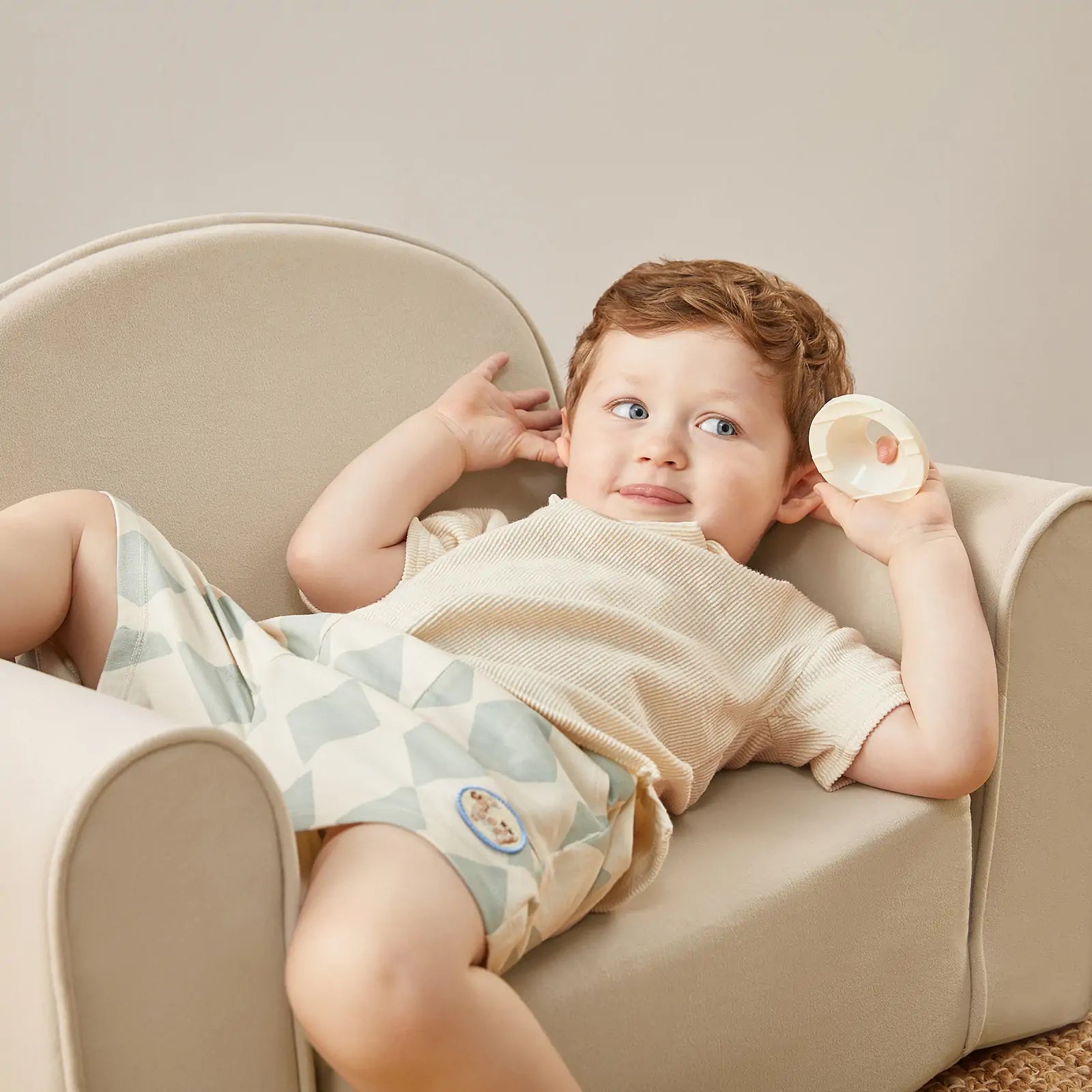 A young child with light brown hair lounges on the Tiny Land® GentleSnug Kid Chair - Beige White by Tiny Land, wearing a light shirt and patterned shorts, playfully holding a round object near their ear.